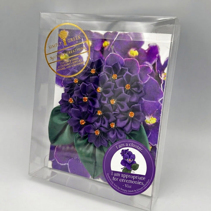 A clear plastic-wrapped purple violet cluster corsage shown on a light gray background. A gold circular sticker reads 'Simply Greek by Utopia Creations' and a purple circular sticker says 'I am a cluster: I am appropriate for ceremonies - Vivi.
