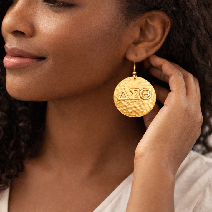 Woman wearing a gold hammered disc Delta Sigma Theta earring with a hand near her ear